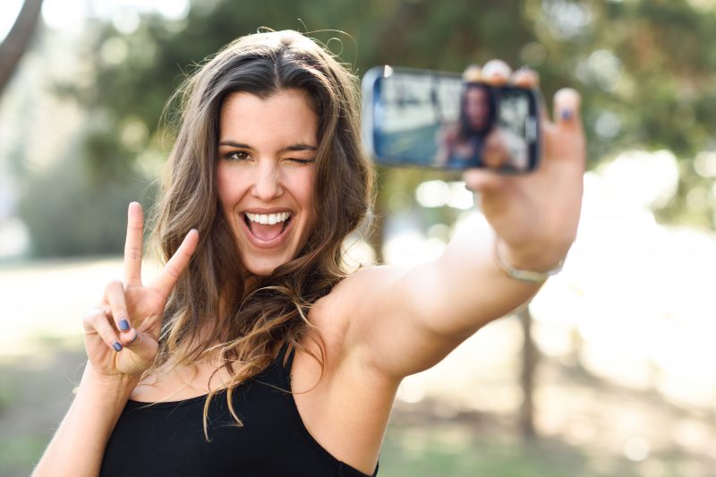 Portrait of a beautiful young woman selfie in the park with a smartphone doing v sign